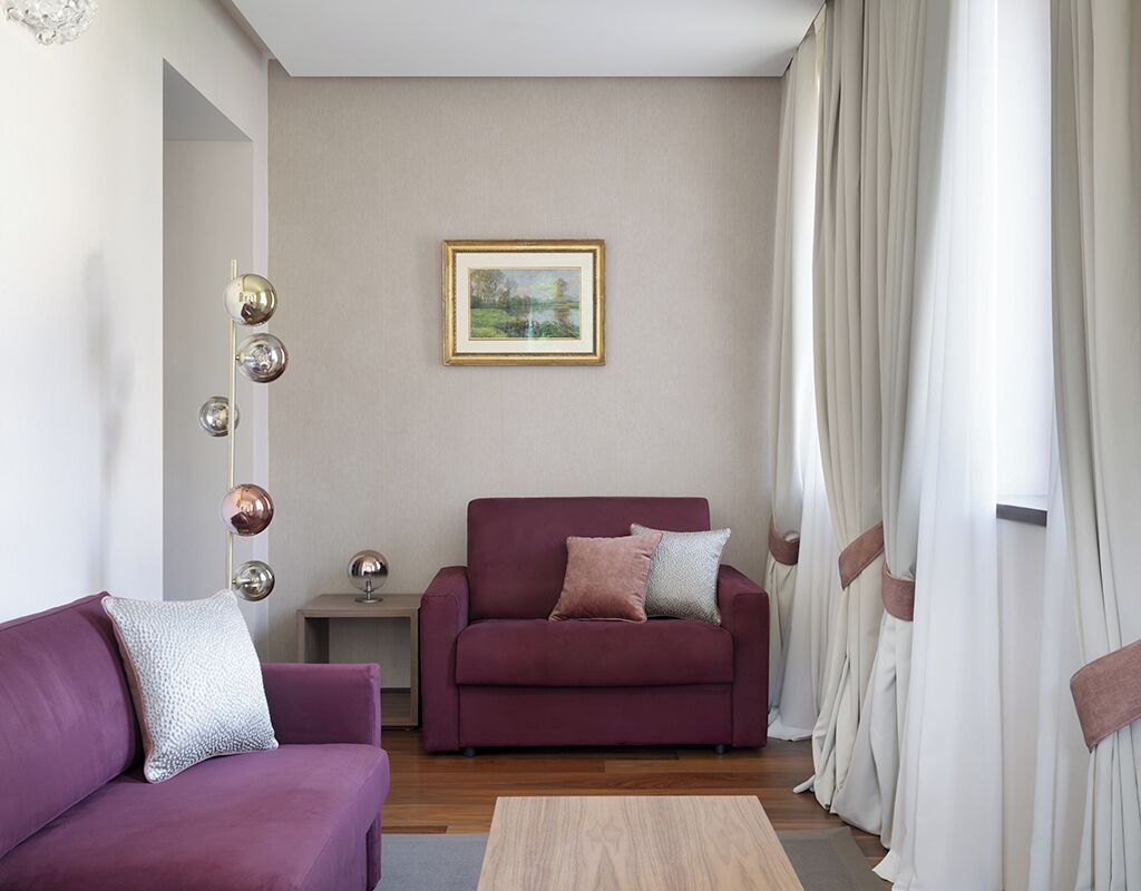 Ein Wohnzimmer mit einem violetten Sofa, einem roten Sessel und einem kleinen Tisch mit einer Lampe. An der Wand hängt ein gerahmtes Bild und es gibt ein Fenster mit Vorhängen.