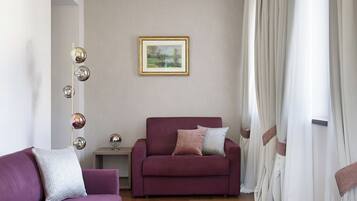 Ein Wohnzimmer mit einem violetten Sofa, einem roten Sessel und einem kleinen Tisch mit einer Lampe. An der Wand hängt ein gerahmtes Bild und es gibt ein Fenster mit Vorhängen.