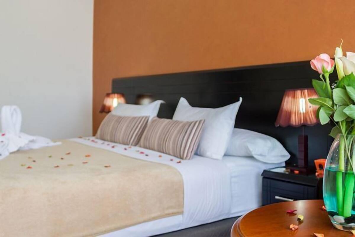 A neatly made bed with white linens and a beige throw, a bedside table with a vase of flowers, and a lamp on the nightstand.