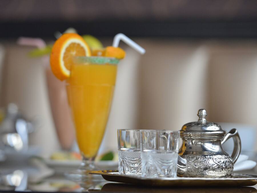 Una mesa de comedor con un vaso alto de jugo de naranja, una tetera plateada y dos vasos pequeños.