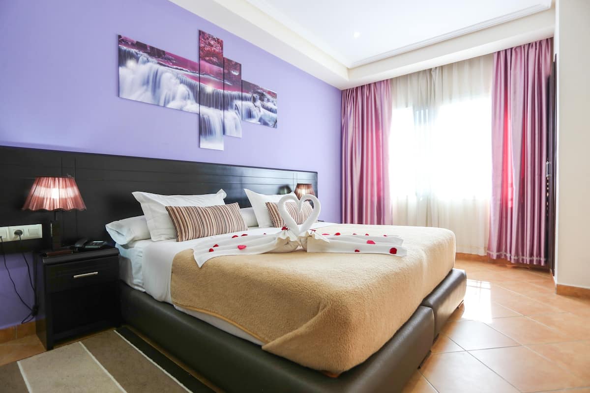 A neatly made bed with a heart-shaped decoration, a bedside lamp, and a wall with abstract art.