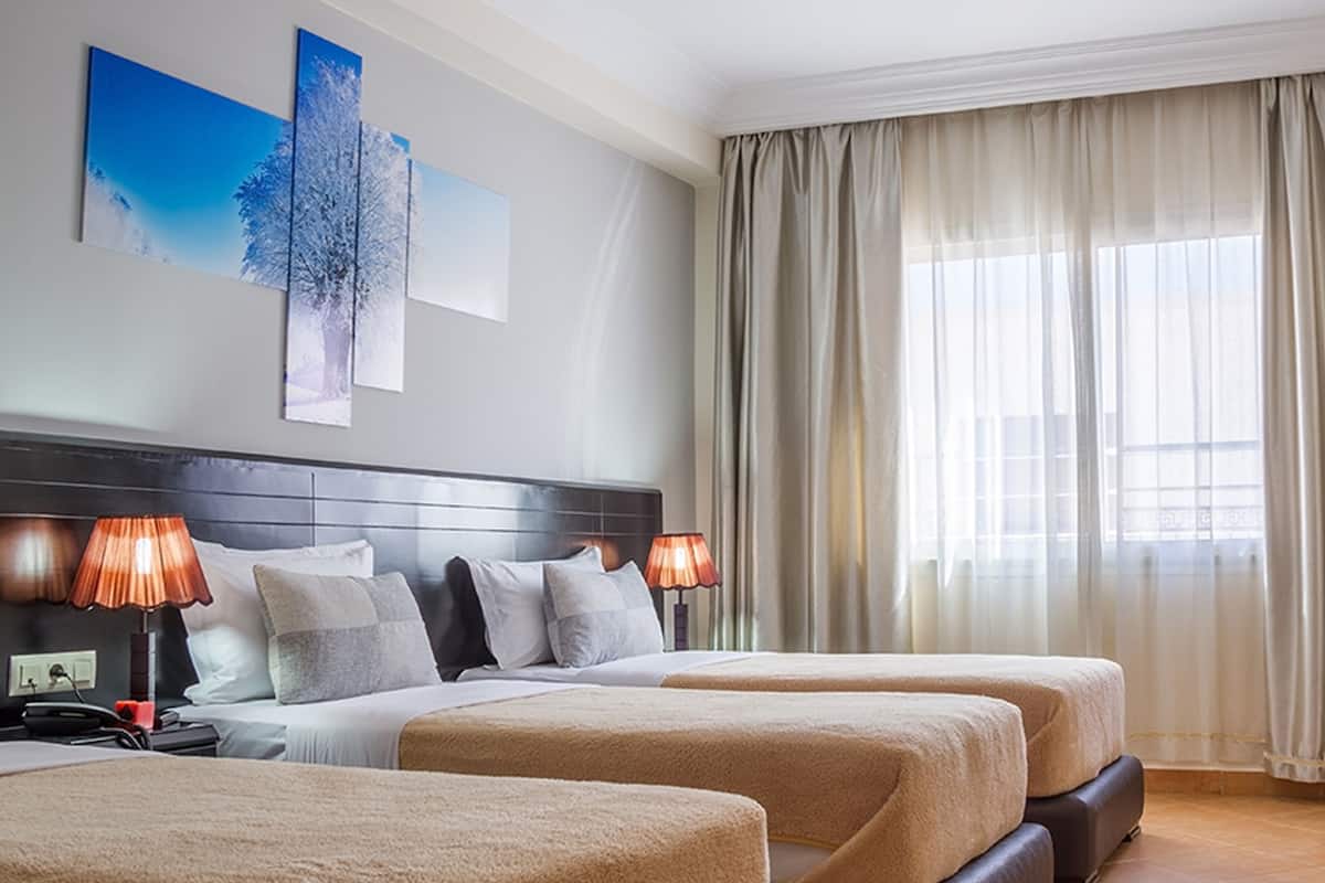 A hotel room with two beds, a large window with sheer curtains, a headboard with a flat-panel artwork, and a bedside table with a lamp.