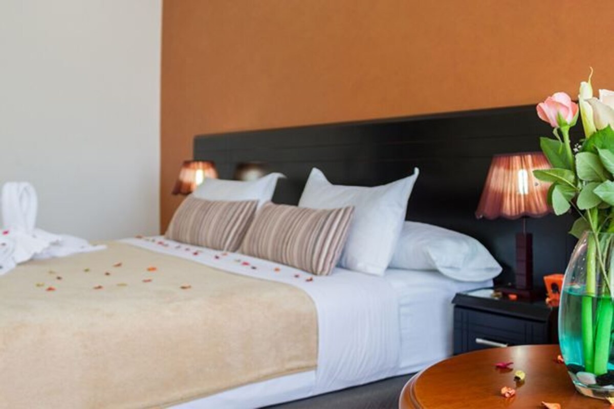 Een netjes opgemaakt bed met witte lakens en een beige plaid, een nachtkastje met een bloevas, en een lamp op de nachtkast.
