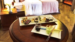 Una mesa de comedor ordenada con variado sushi y un tazón pequeño de salsa de soja, frente a un dormitorio elegante con una cama y una chimenea.
