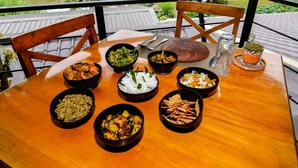 Una mesa de madera con varios platos, incluyendo arroz, verduras y pan, ubicada en un balcón con vista a un lago y vegetación.