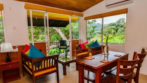 Comedor con muebles de madera, una mesa de vidrio y ventanas amplias que dan a vista de árboles y un lago.