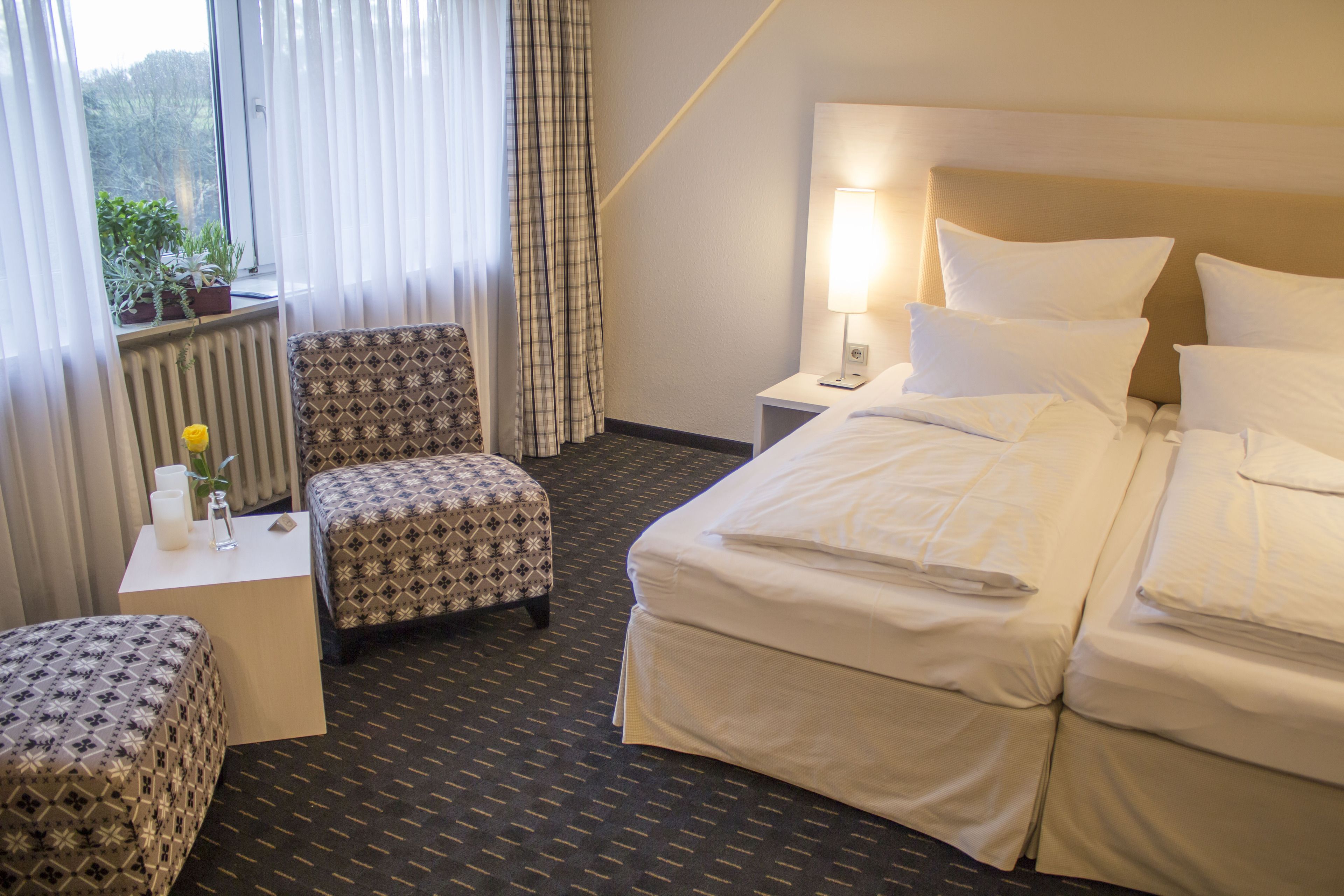 A hotel room with a bed, a chair, a small table with a lamp, and a window with curtains.