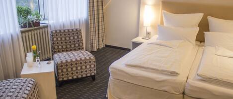 A hotel room with a bed, a chair, a small table with a lamp, and a window with curtains.