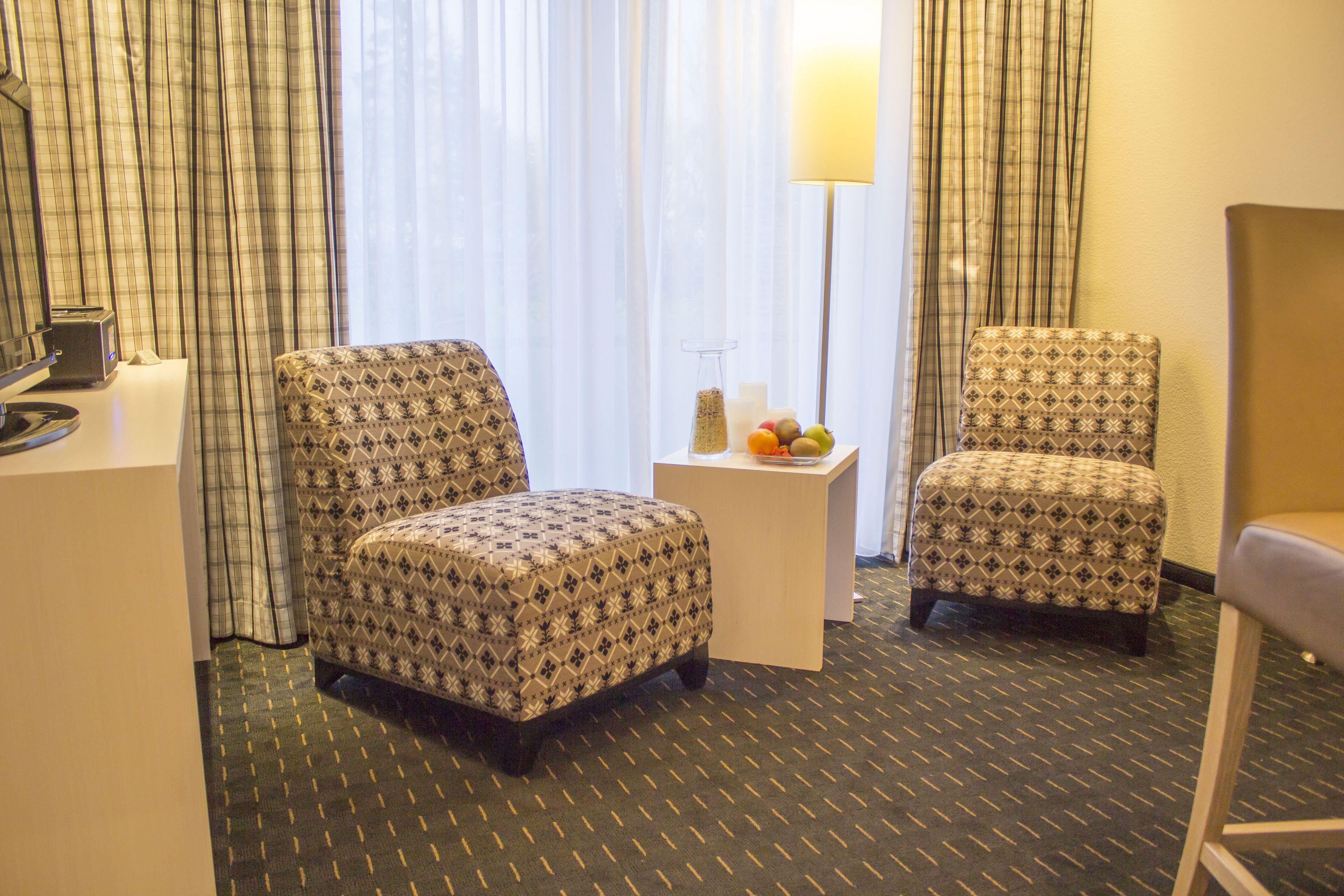 Habitación de hotel con dos sillones estampados, una mesita con lámpara y vistas al exterior a través de cortinas traslúcidas.