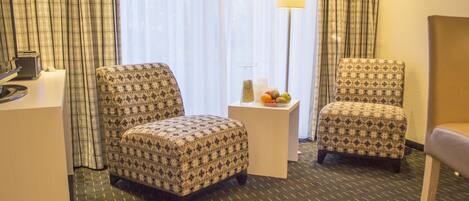 Habitación de hotel con dos sillones estampados, una mesita con lámpara y vistas al exterior a través de cortinas traslúcidas.