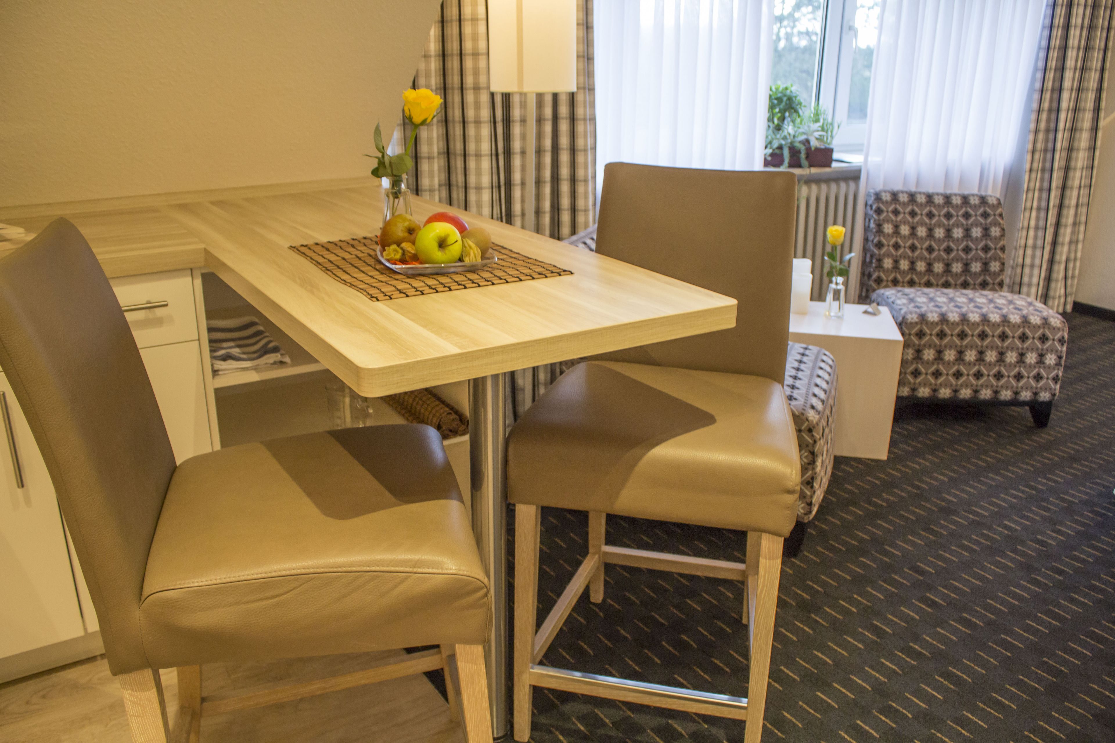 A hotel room with a wooden dining table, two leather chairs, a patterned armchair, a small side table, and a window with curtains.