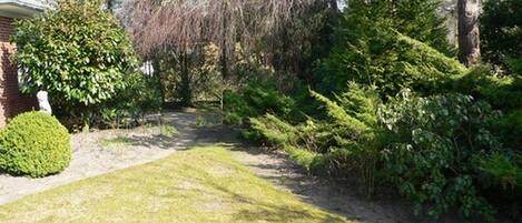 A well-maintained garden with a variety of trees and shrubs, a paved path, and a brick building in the background.