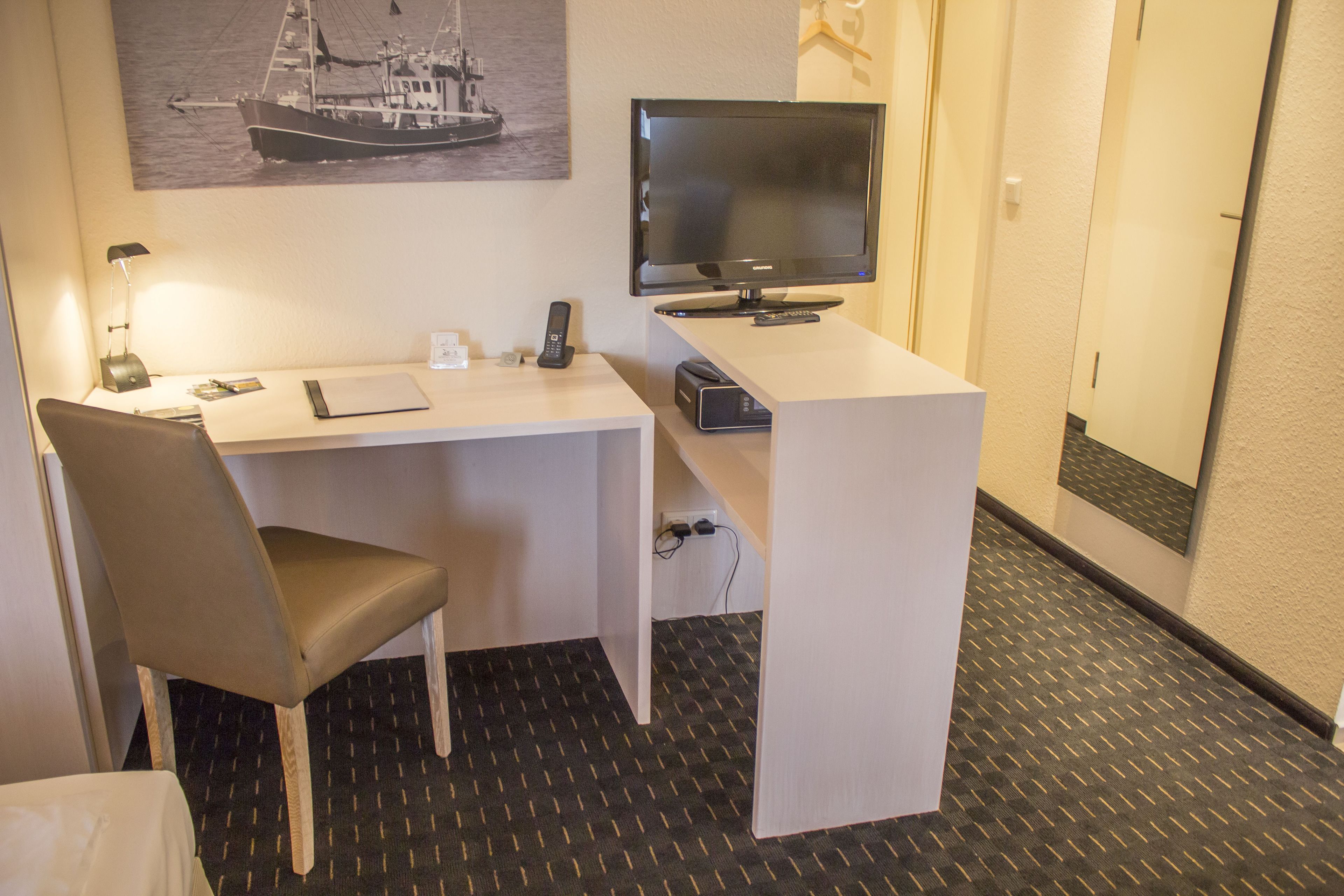 A hotel room with a desk, chair, television, and a painting of a boat on the wall.