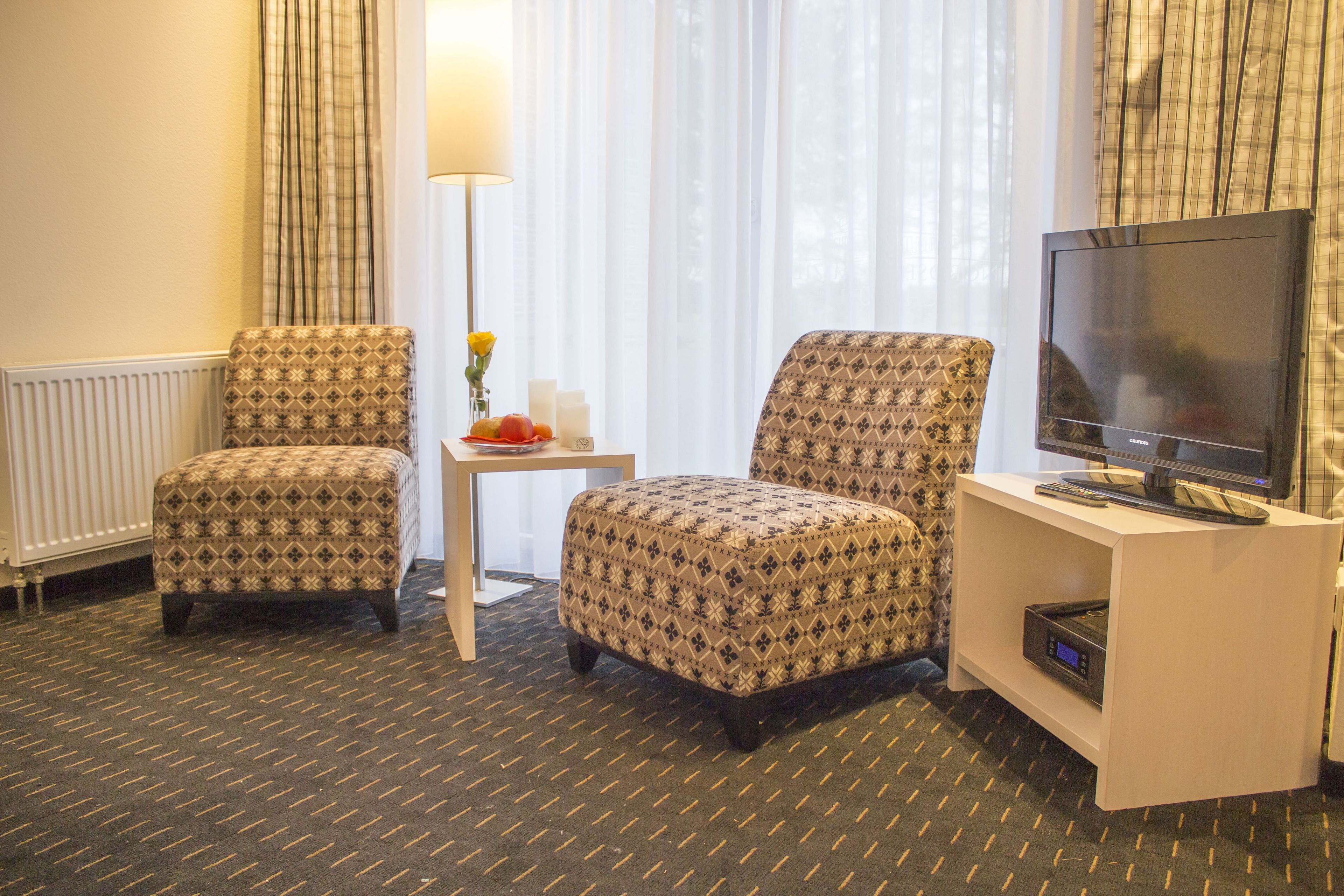 Habitación de hotel con dos sillones estampados, una mesita con un florero y fruta, un televisor de pantalla plana y un radiador.