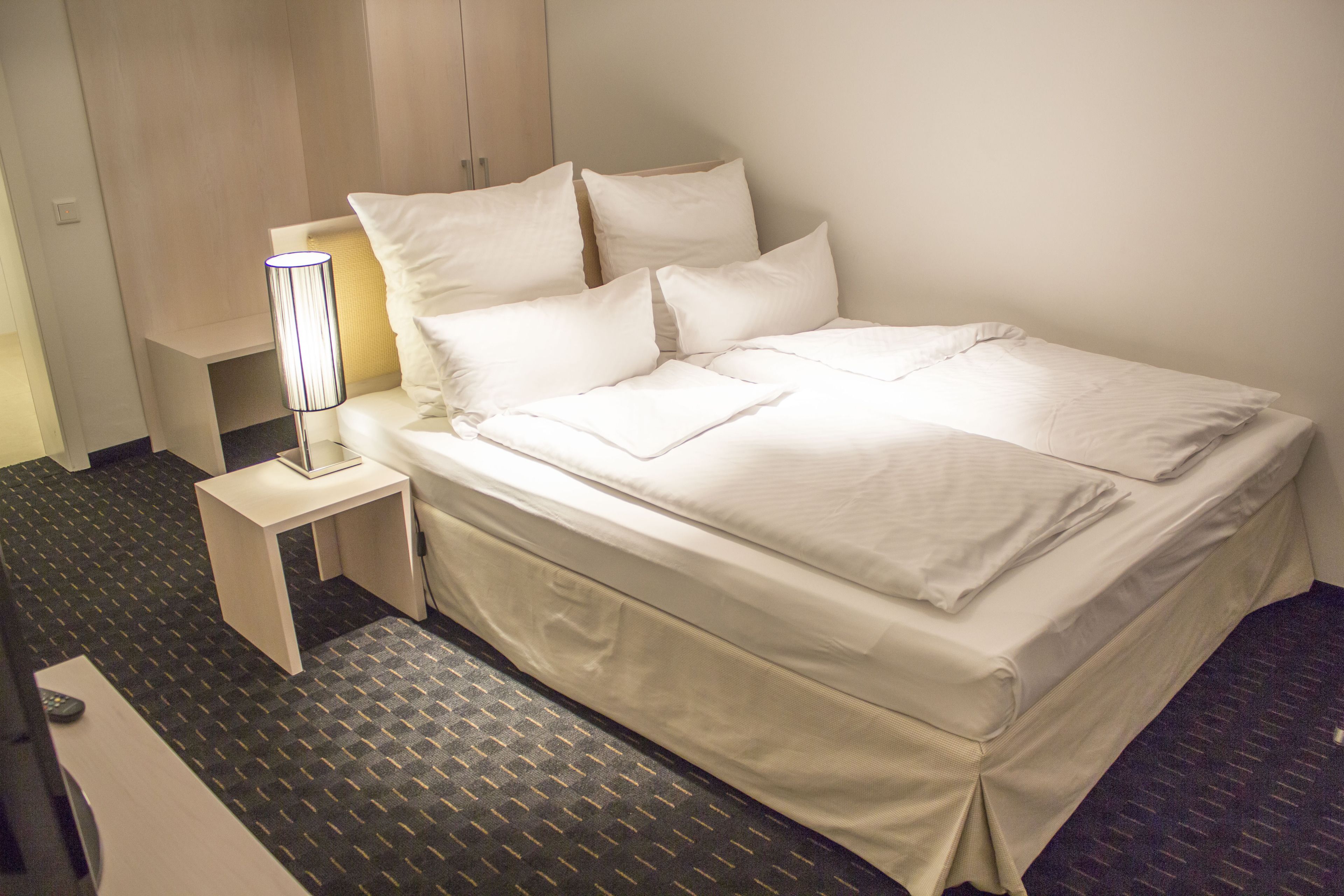 A neatly made bed with white linens, a bedside table with a lamp, and a carpeted floor.