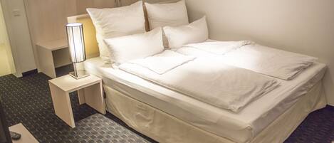 A neatly made bed with white linens, a bedside table with a lamp, and a carpeted floor.