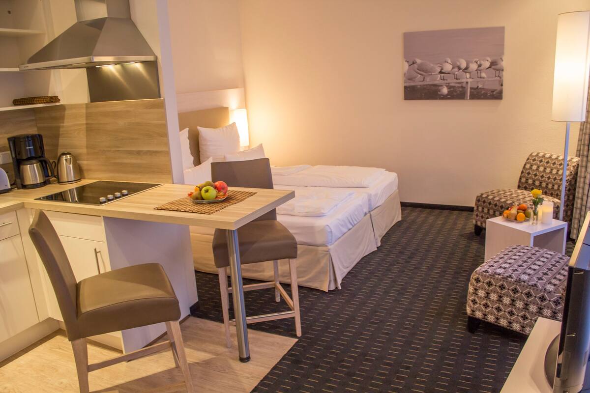 Een compacte hotelkamer met een kitchenette, een bed, een klein eettafeltje met fruit, een gestreept fauteuil en een nachtkastje met een lamp.
