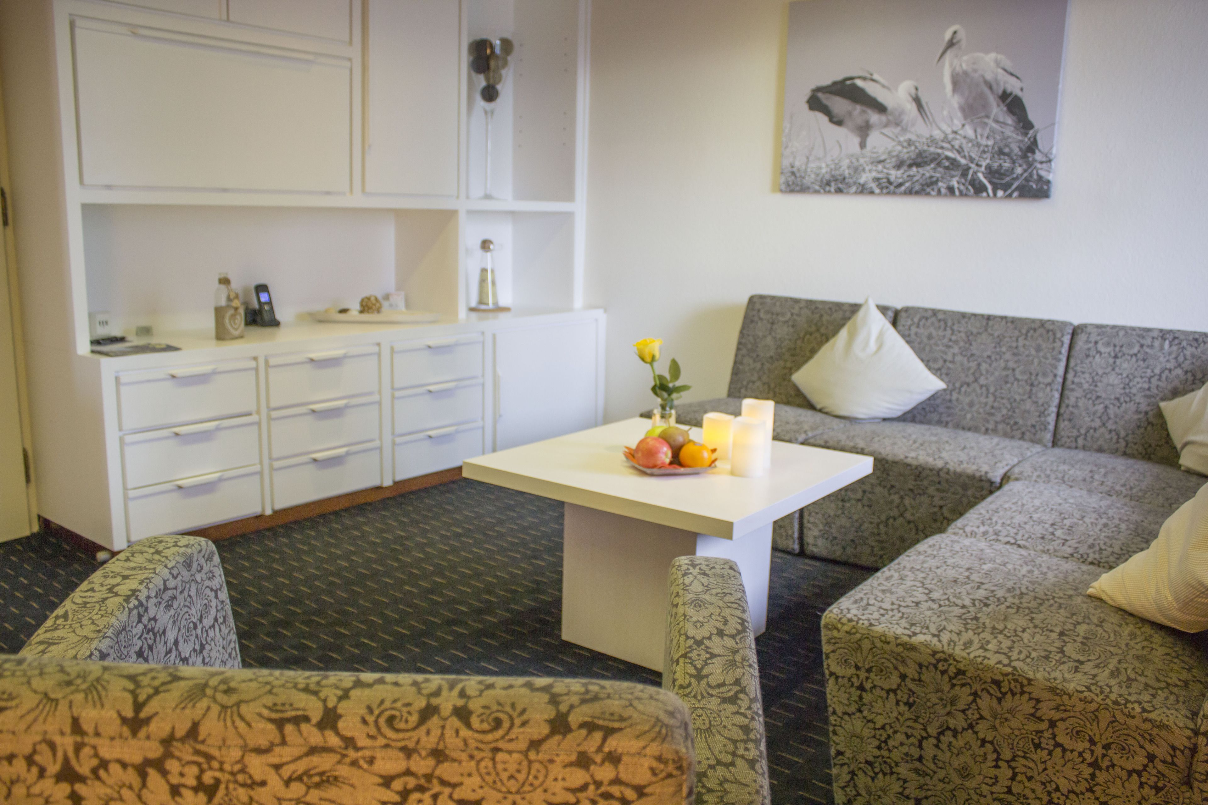 Una habitación de hotel moderna con un sofá de color gris con estampado, una mesa blanca con fruta y velas, un tocador blanco y un cuadro enmarcado en la pared.