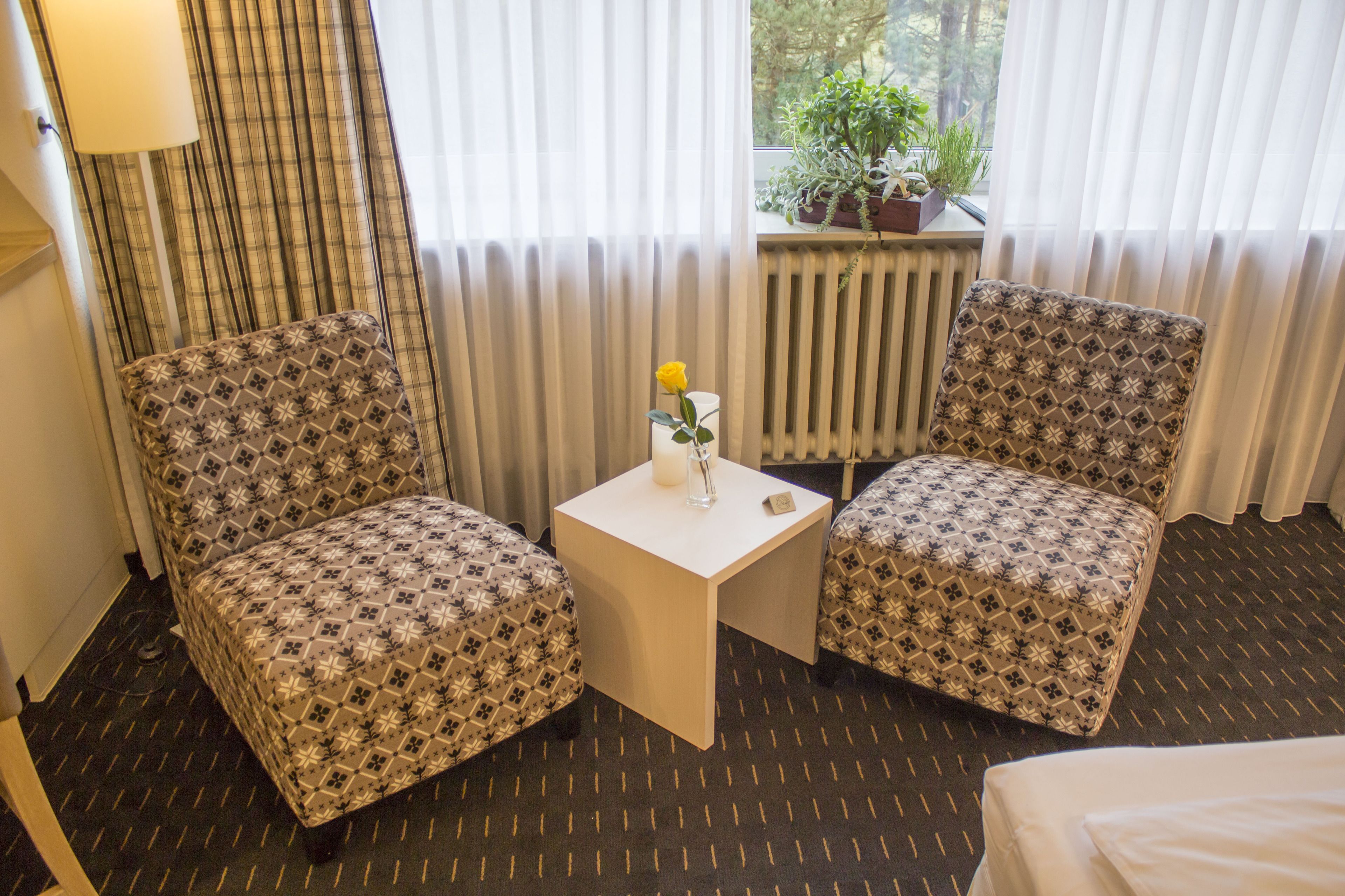 A hotel room with two patterned armchairs, a small table with a candle and a vase of flowers, a radiator, and a window with a view of greenery.