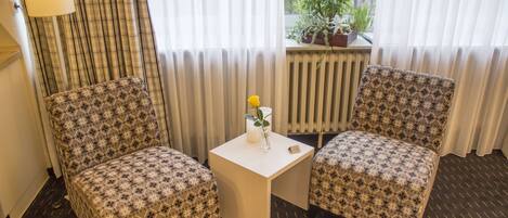 A hotel room with two patterned armchairs, a small table with a candle and a vase of flowers, a radiator, and a window with a view of greenery.