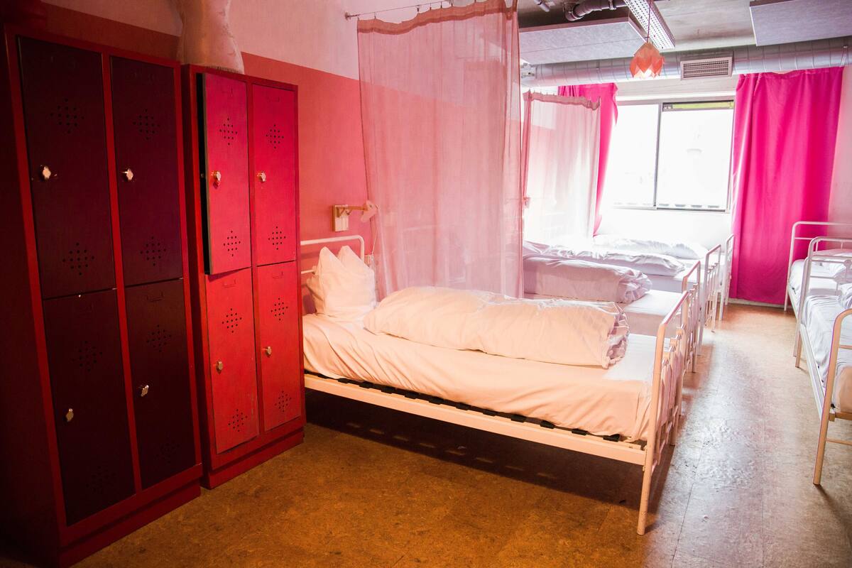 A room with a single bed, a window with pink curtains, and a row of red lockers.