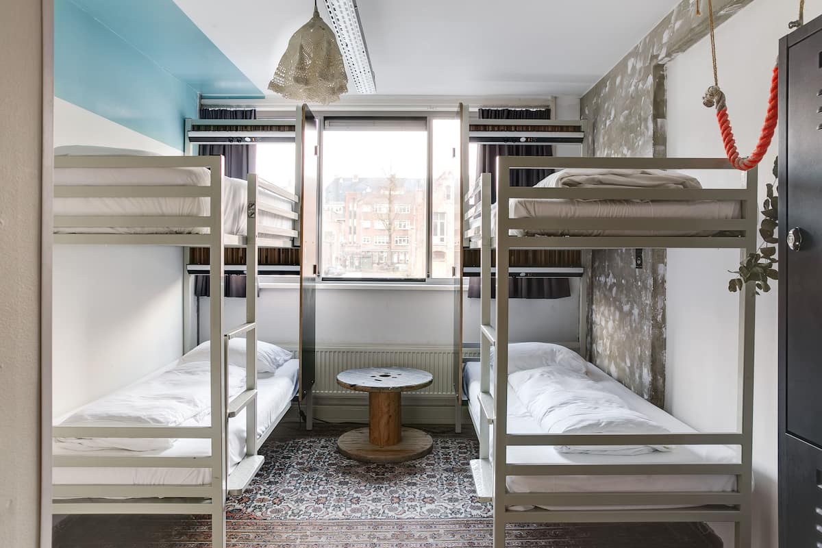 A dormitory room with bunk beds, a small round table, and a view of buildings through a window.