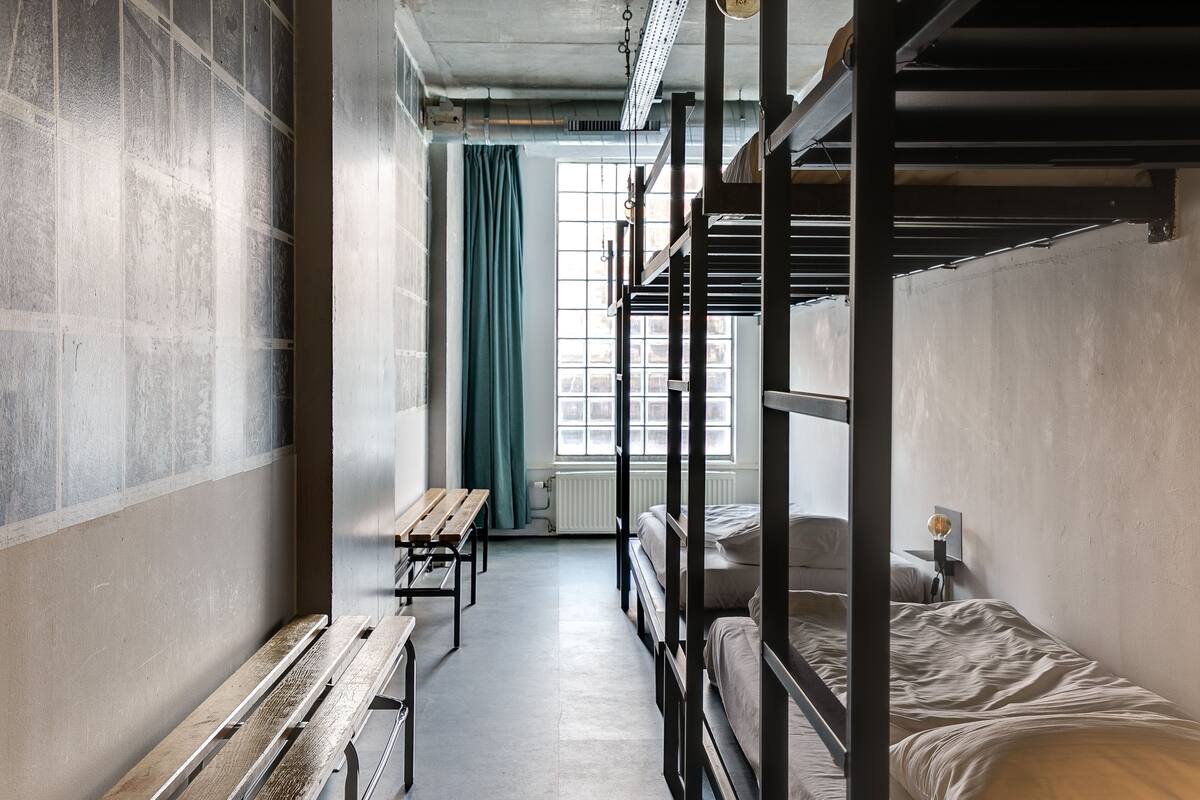 A dormitory room with bunk beds, a wooden bench, and a window with a curtain.