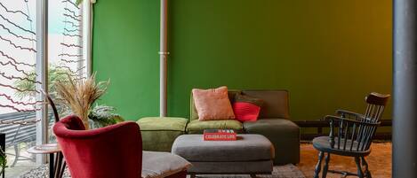 A cozy seating area with a red chair, grey ottoman, and a black chair. A green wall and a window with blinds are visible.