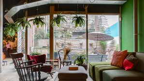 A cozy living room with a green sofa, a wooden chair, and a small table with a book. There are potted plants and a view of the outside through large windows. - ClinkMama (Amsterdam)