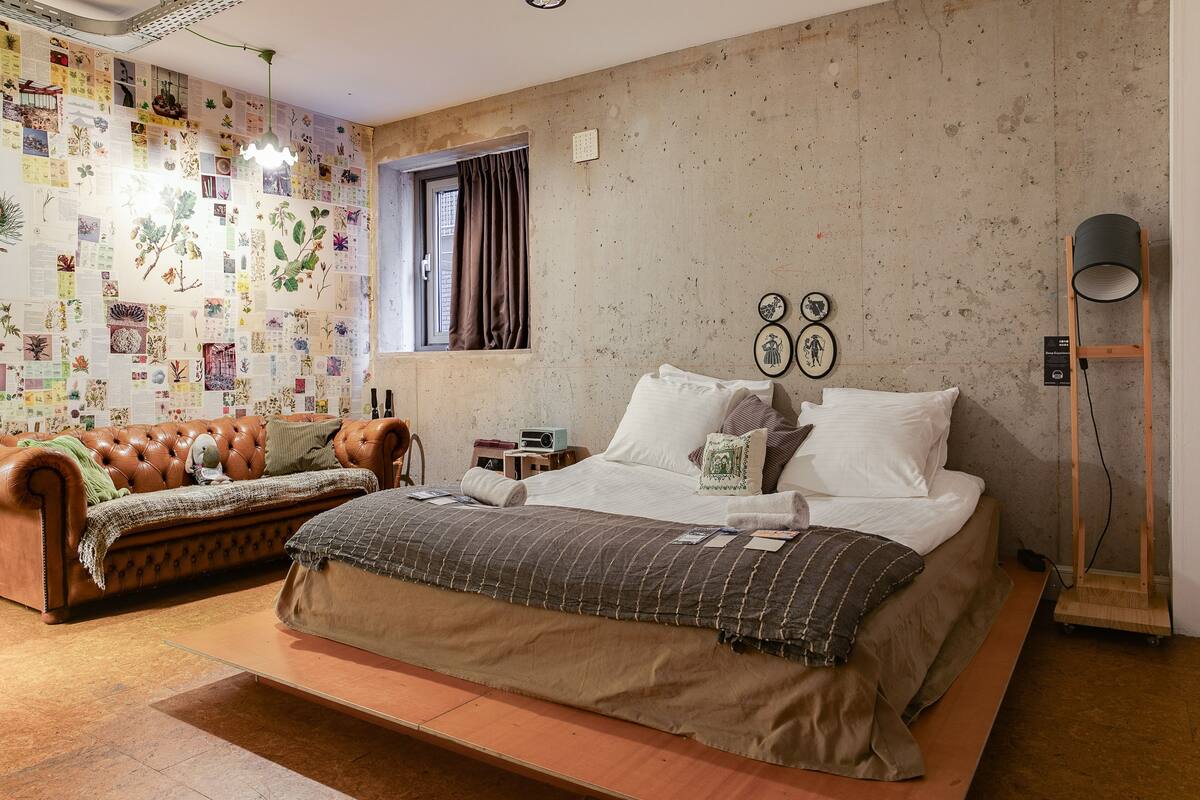 A bedroom with a bed, a sofa, a small table, a lamp, and a wall with botanical illustrations.