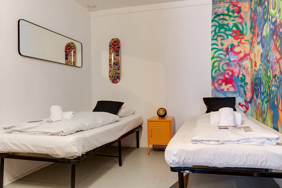 A room with two beds, a mirror, a colorful graffiti wall, and a small yellow cabinet.
