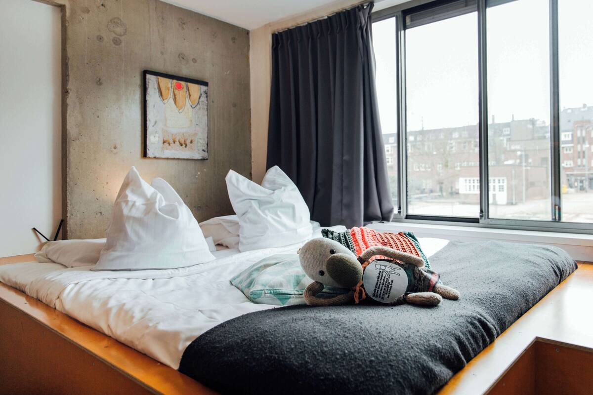 A hotel room with a bed, pillows, a stuffed toy, a window with a view of buildings, and a wall with a map.