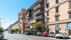 Vista de la calle con una fila de coches aparcados, una línea de tranvía y un edificio de estilo clásico con varios balcones.