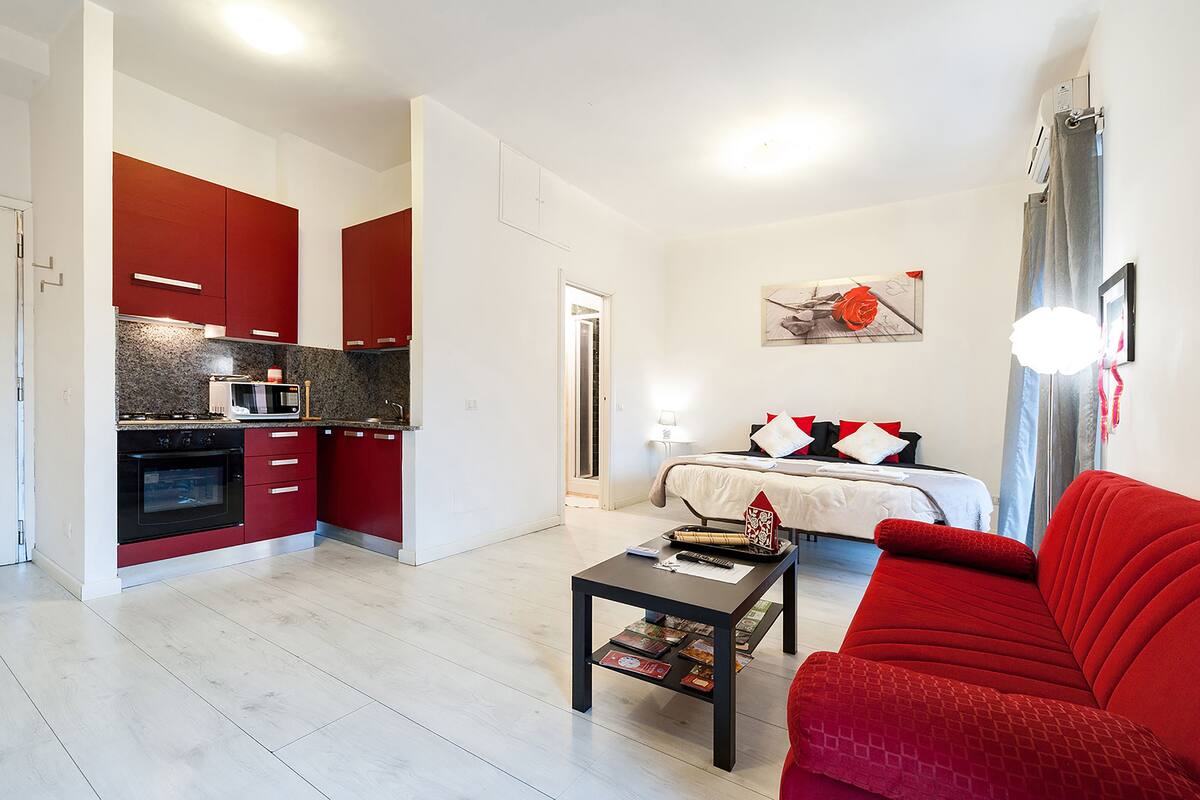 Un espacio habitable compacto con kitchenette, una cama con ropa de cama roja y blanca, un sofá rojo y una mesa de centro con libros.