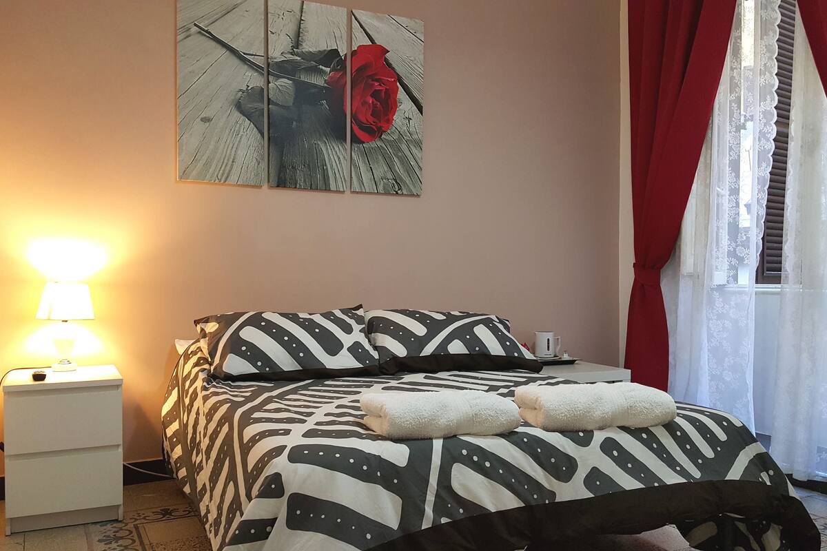 Un dormitorio con una cama, una mesita de noche, una lámpara, un cuadro de una rosa y una ventana con cortinas.