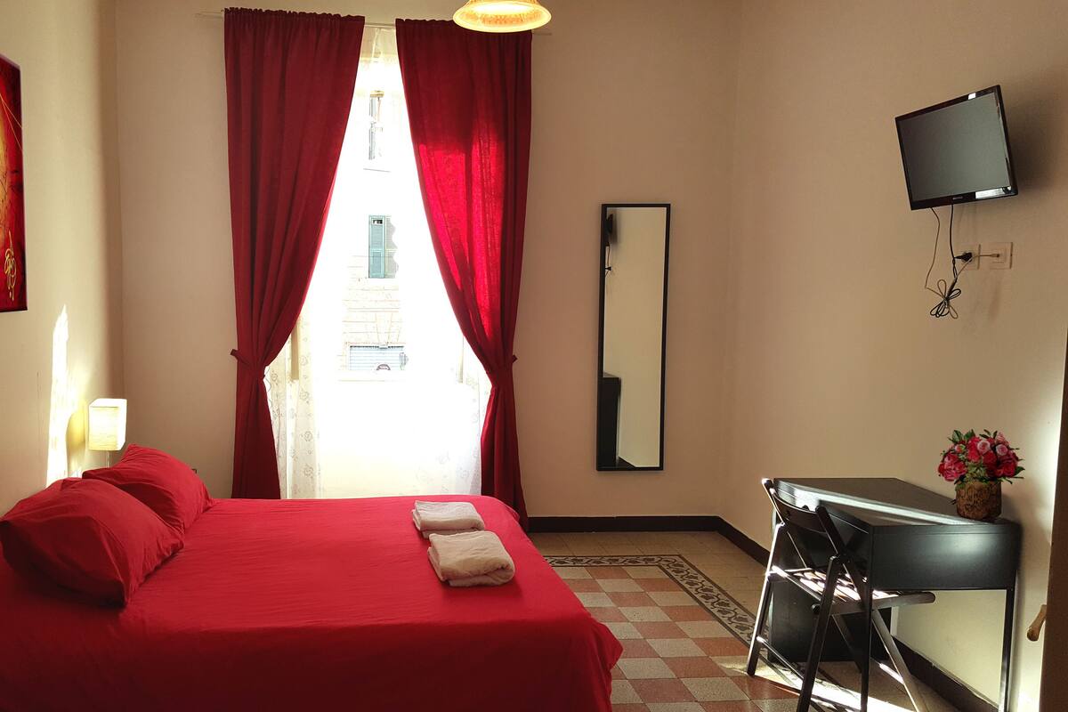 Una habitación de hotel con una cama roja, un escritorio con silla, un televisor y un espejo.