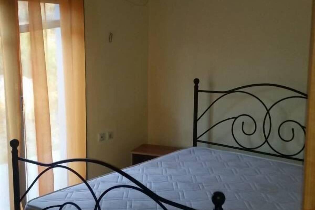 Un lit simple avec un cadre en métal et un matelas blanc, dans une pièce dont le mur est uni et qui possède une fenêtre avec des rideaux.