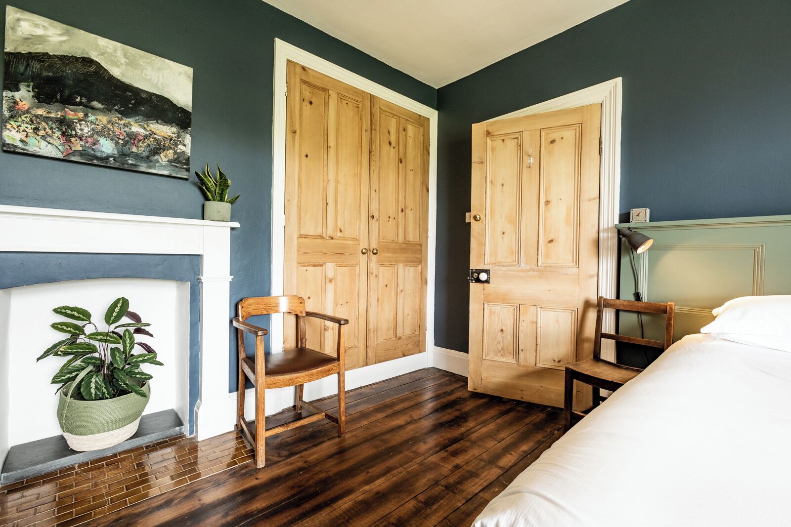 Une chambre avec des murs sombres, un plancher en bois, deux portes en bois, un lit blanc, une chaise, une plante en pot et un tableau au-dessus de la cheminée.
