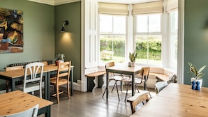 A dining area with a bay window, wooden tables and chairs, and a potted plant. - The Old Vicarage (Cardigan)