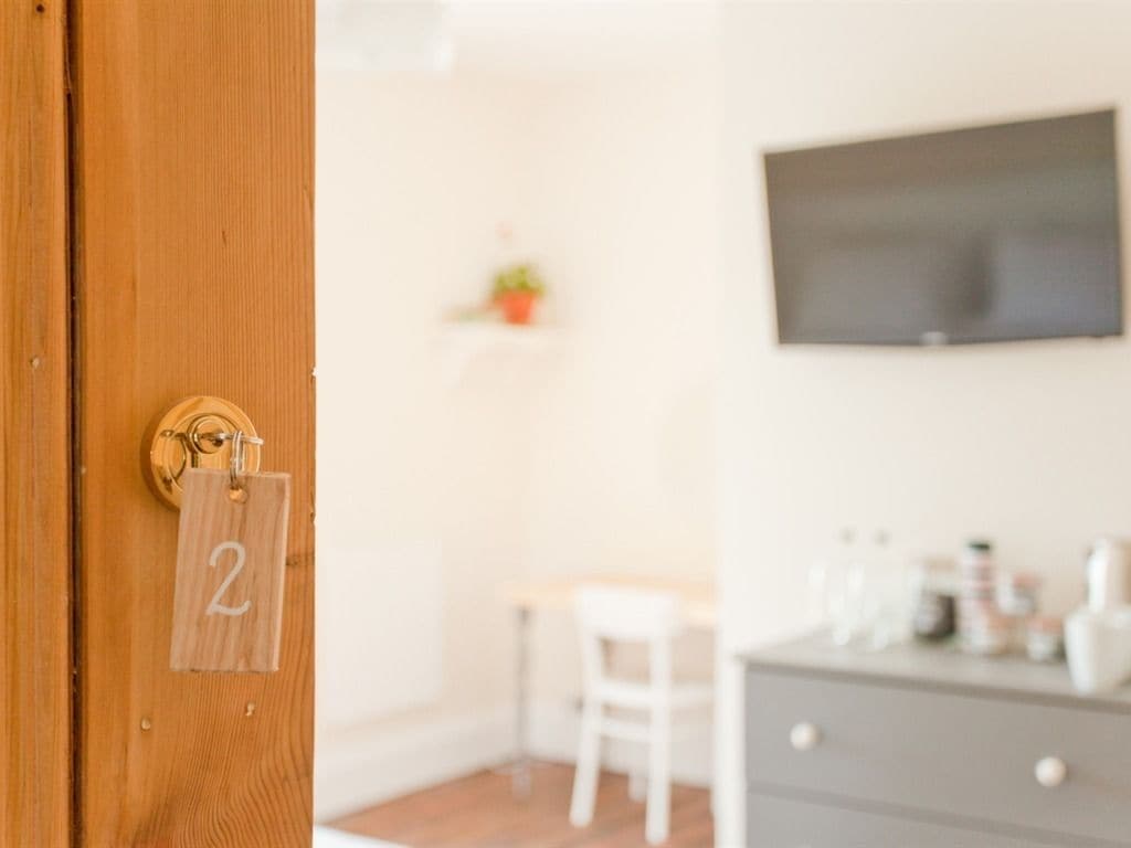 Una habitación con una puerta de madera que indica el número 2, una silla blanca, una mesita pequeña, un aparador gris y un televisor montado en la pared.