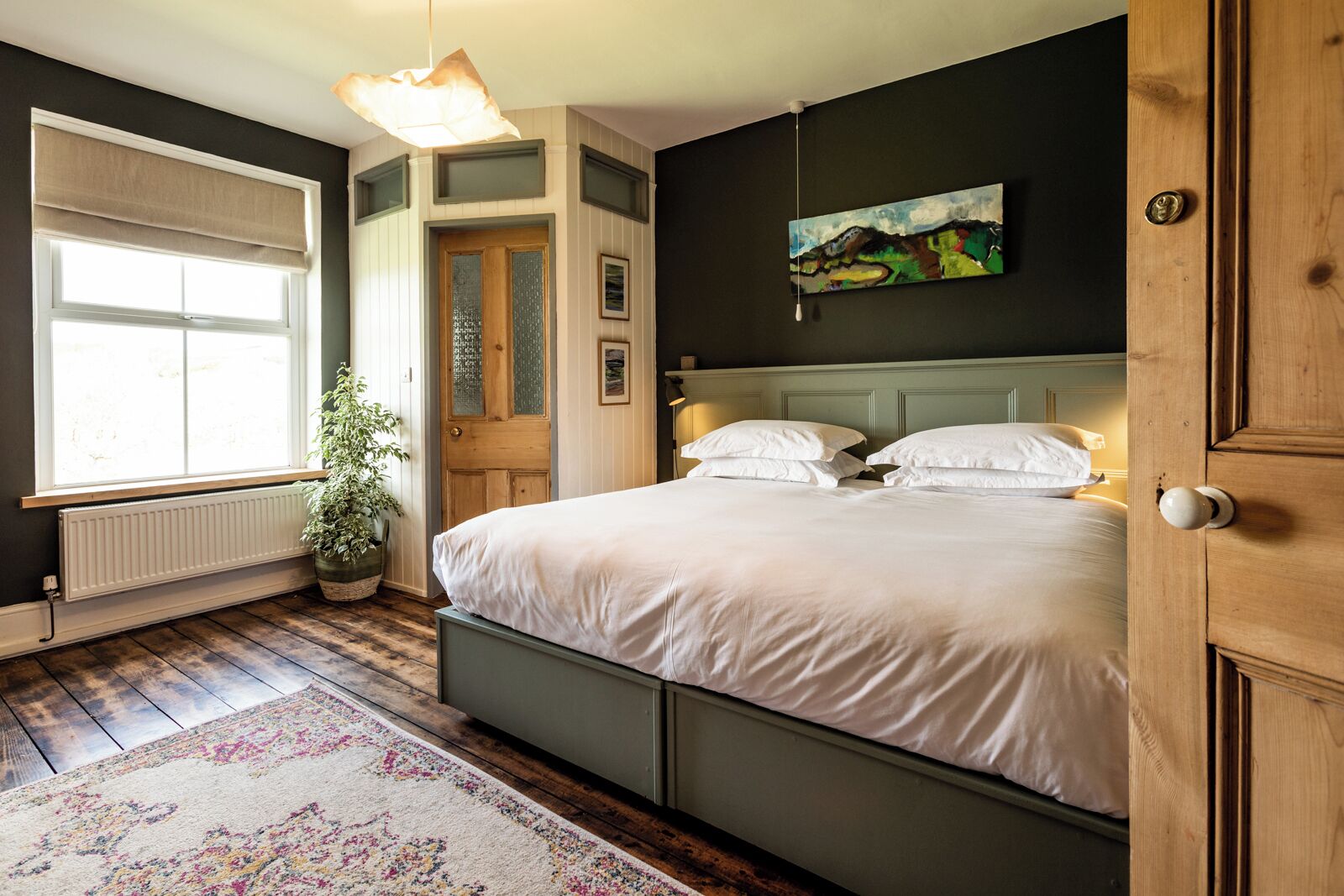 Un dormitorio con una cama grande, una ventana con persianas, un radiador, una planta en maceta y un cuadro en la pared.