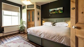 Un dormitorio con una cama grande, una ventana con persianas, un radiador, una planta en maceta y un cuadro en la pared.