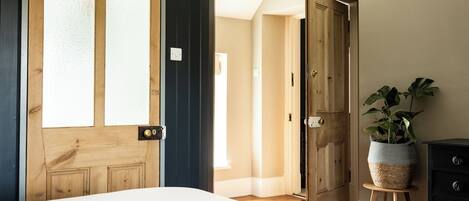 A bedroom with a large window, a wooden door, a bedside table with a plant, and a dresser.