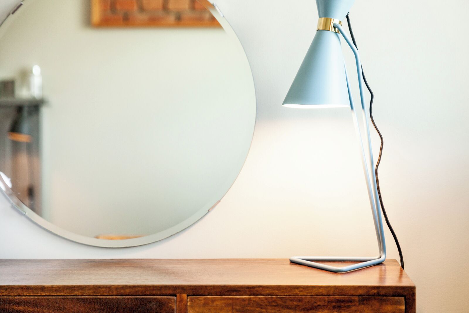 A round mirror on a wooden dresser with a blue desk lamp beside it.