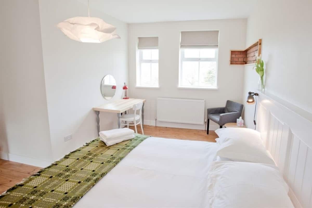 A bedroom with a bed, a desk, a chair, a mirror, and two windows.