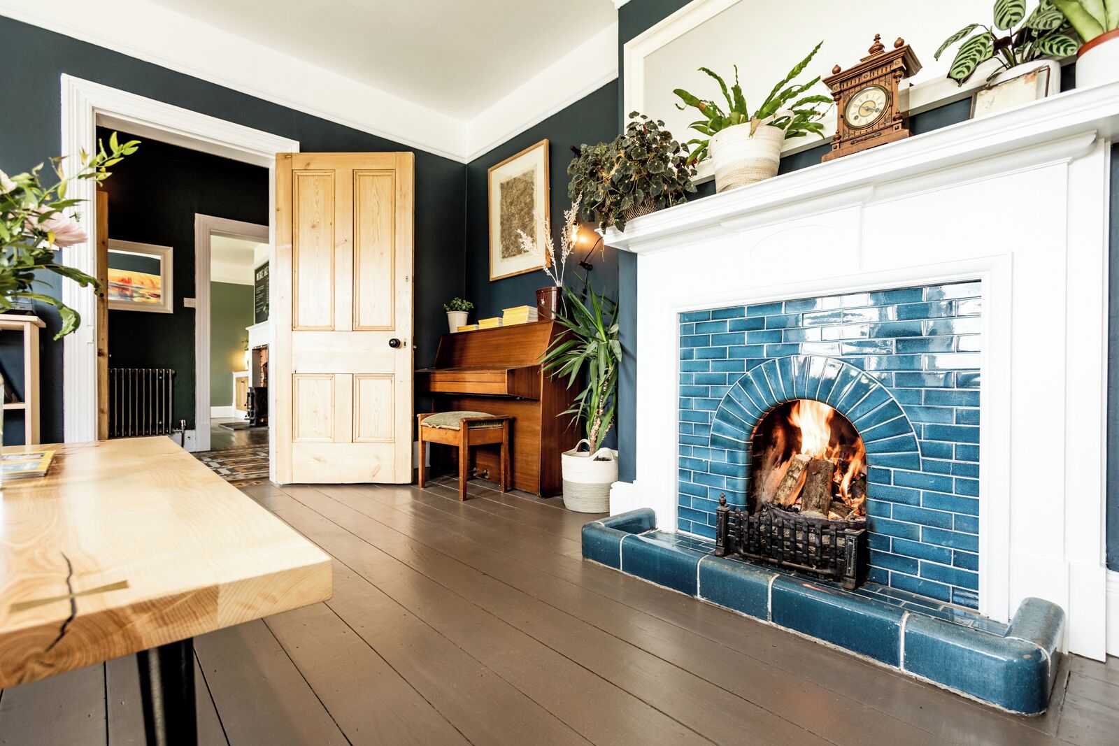 Een gezellige woonkamer met een blauw betegelde open haard, een houten piano en een deur naar een andere kamer.