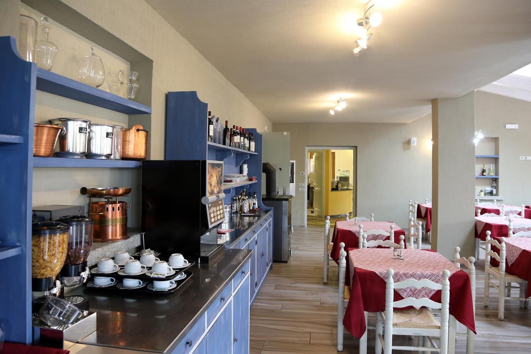 A dining area with tables covered in red tablecloths, a kitchen area with copper pots and a coffee machine, and a hallway leading to another room.
