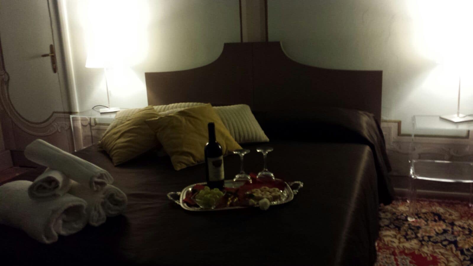 A hotel room with a neatly made bed, a tray with wine and fruit, and towels on the side.