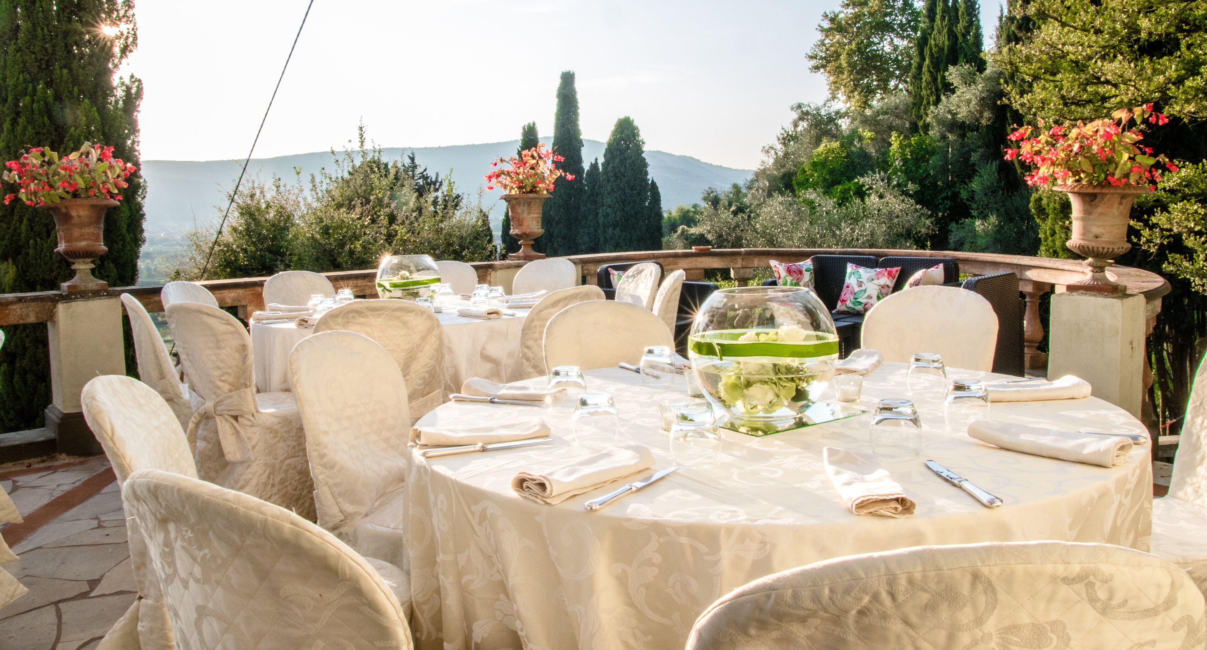 Una mesa redonda con manteles blancos y sillas, un cuenco de vidrio con flores, y una vista panorámica de árboles y montañas al fondo.