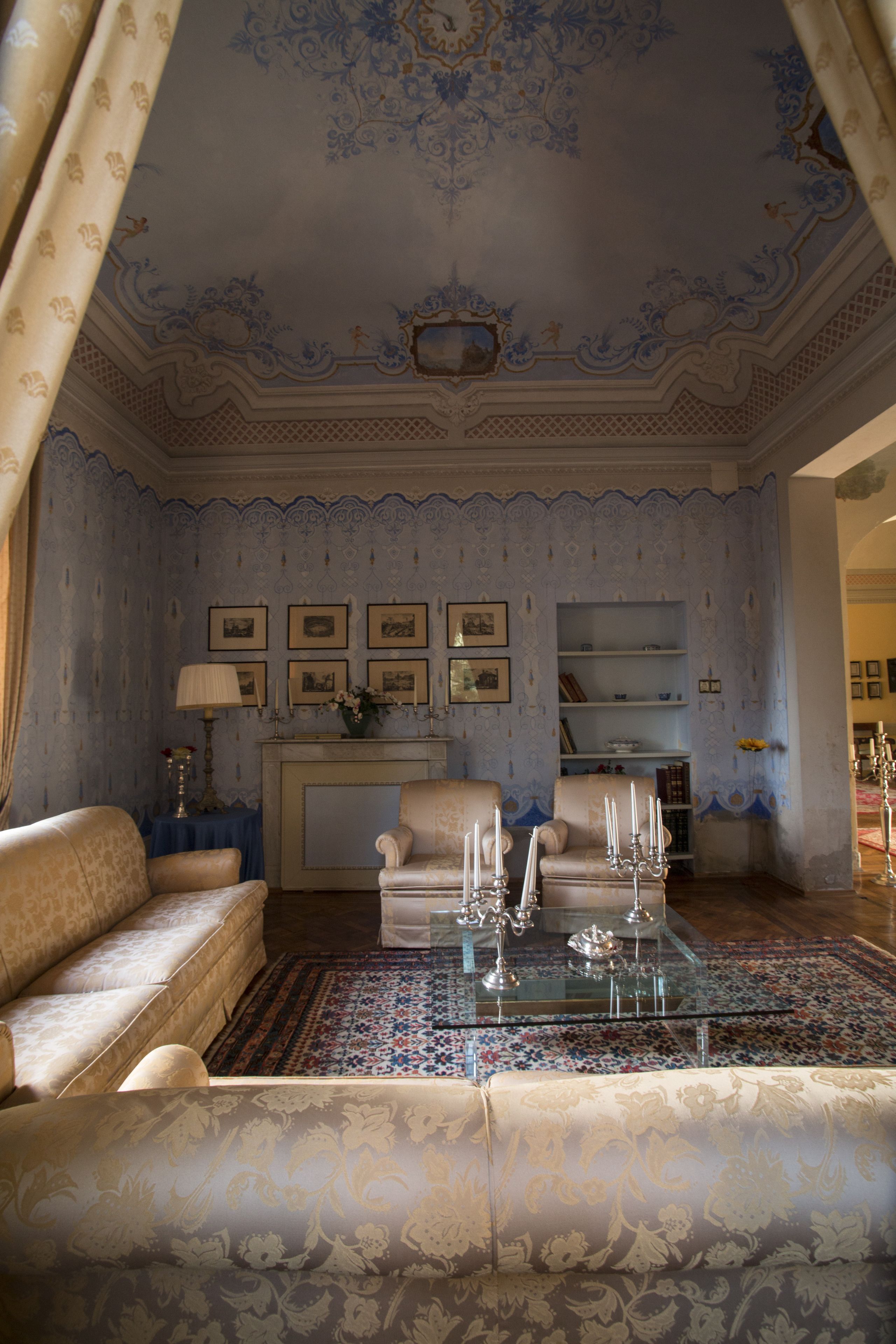 A room with a patterned wall, a sofa, a chair, a glass coffee table, and a fireplace.